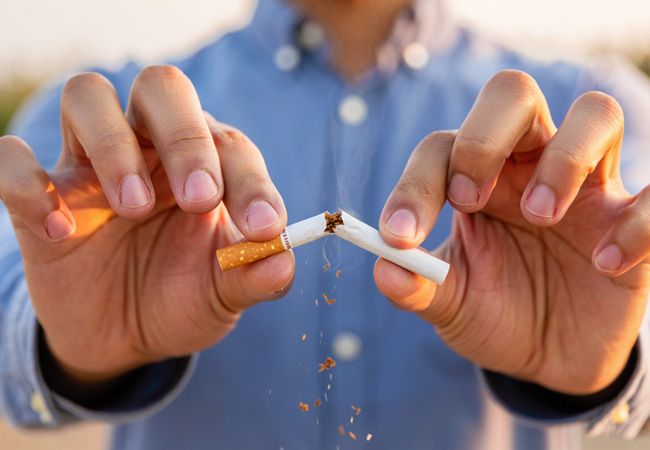 Мужские руки в синей рубашке ломают сигарету пополам на закате